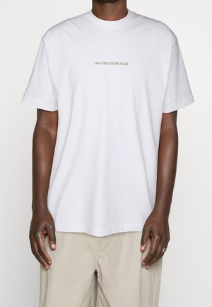Osoba w białej koszulce z krótkim rękawem z napisem "ON VACATION CLUB" w małej zielonej czcionce na klatce piersiowej, w połączeniu z beżowymi spodniami.