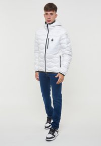 Hvid pufferjakke med quiltet design, der har hætte, frontlynlås og en sort brystlomme. Stylings med blå jeans og sorte sneakers.