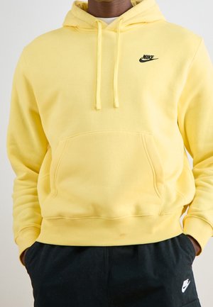Persoon die een gele Nike-hoodie draagt met voorkantzak en trekkoorden, gecombineerd met zwarte Nike-broek met logo op het linkerbeen.