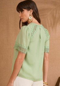 Blusa verde menta con maniche corte a sbuffo, caratterizzata da una delicata finitura in pizzo sulle maniche e sul collo. Tessuto morbido e leggero, dettaglio a chiave sul retro.