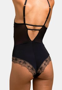 Le body noir présente un tissu en maille transparente, des bretelles ajustables et un bord en dentelle florale en bas, mettant en valeur une finition lisse et une silhouette définie.