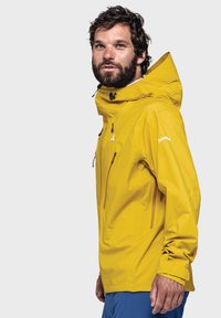 Gelbe wasserdichte Jacke mit hohem Kragen, verstellbarer Kapuze, praktischen Reißverschluss-Taschen und elastischen Bündchen. Glatte Textur mit dezenten Mustern.