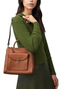Sac à main en cuir marron avec une poche avant à rabat et des ferrures dorées, tenu par une personne portant un pull texturé vert.