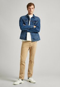 Chaqueta de mezclilla azul con dos bolsillos frontales, sobre una camisa blanca; pantalones beige; zapatillas blancas con acentos verdes; de pie con los brazos cruzados.