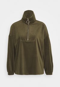 Suéter de pana verde oliva con un alto cuello, frontal con cremallera y un anillo metálico prominente como tirador. Presenta mangas holgadas y una textura característica.
