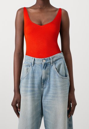 Femme portant un haut sans manches côtelé rouge vif et un jean en denim bleu clair taille haute sur fond uni.