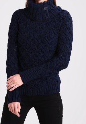 Femme portant un pull en maille bleu marine avec un col montant et trois boutons, posant les bras croisés sur un pantalon noir.