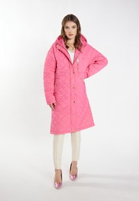 Cămașă lungă roz matlasată cu glugă, având accesorii aurii, un buzunar cu fermoar și închidere cu buton prin presiune. Purtați-o peste pantaloni crem și tocuri roz strălucitoare.