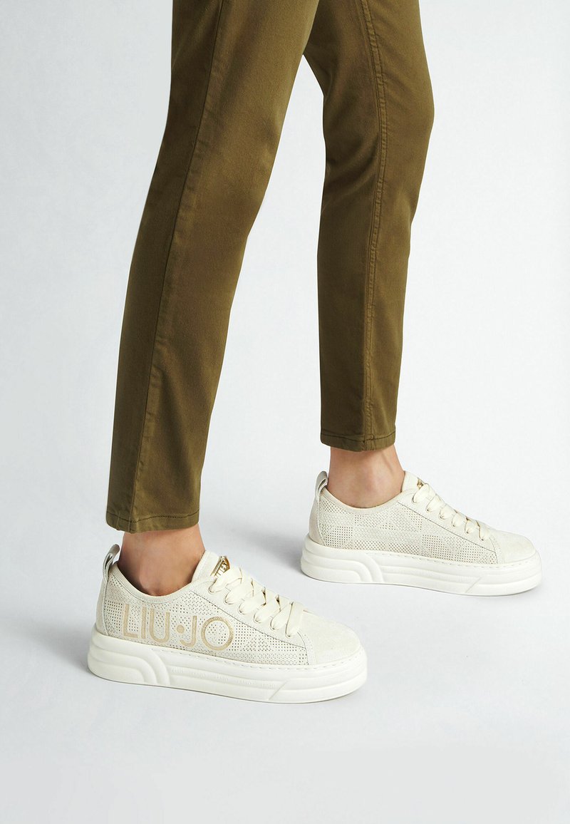 Sneaker bianchi con texture, pannelli in rete e suola piatta. Logo "LIU JO" in oro sul lato. Indossati con pantaloni aderenti verde oliva.