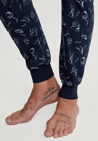 Navy gestreepte pyjamabroek met ski- en bergmotieven. Omgeklapte pijpen en zachte stof. Blootsvoets met zichtbare tattoos.