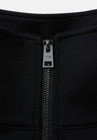 El detalle de tela negra con cremallera cuenta con un tirador de cremallera suave con la marca "MNG" grabada. El material parece elástico y texturizado.