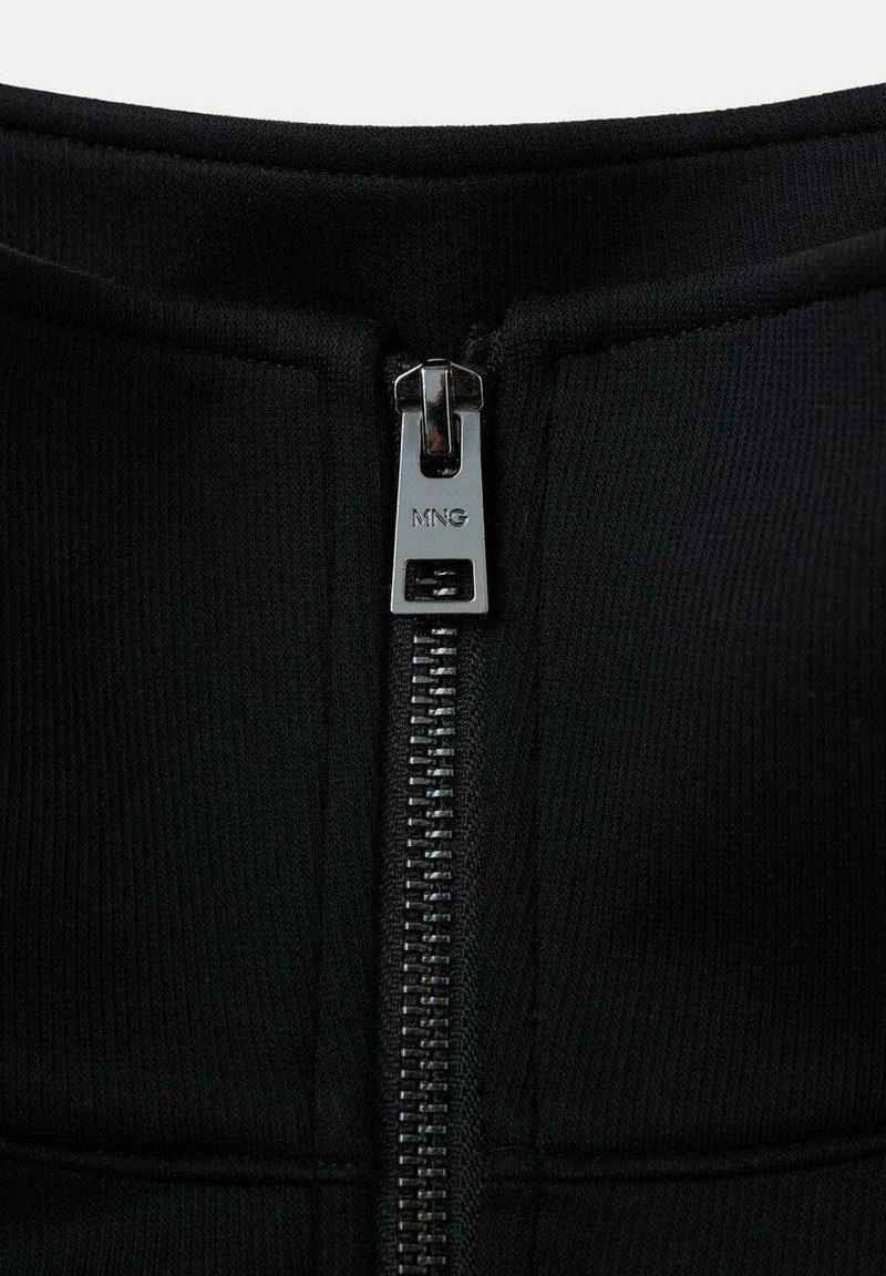 Schwarz, mit Reißverschluss versehenes Stoffdetail, das einen glatten Reißverschlussgriff mit dem eingeprägten Markennamen "MNG" aufweist. Das Material wirkt dehnbar und texturiert.