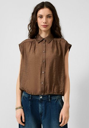Frau mit langen dunklen Haaren, die eine ärmellose braune Bluse mit Knöpfen und blaue Jeans trägt und vor einem schlichten hellen Hintergrund steht.