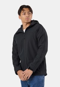 Young man wearing a black hooded jacket and blue jeans, looking to the side with hands clasped in front, against a plain background.