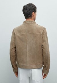 Braune Wildlederjacke mit strukturiertem Kragen, vertikalen Nähten und minimalen Hardwaredetails. Glatte Textur, taillierte Passform und hüftlanger Schnitt.