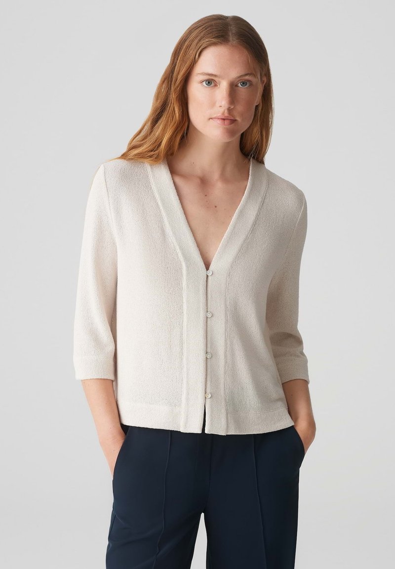 Hellbeige Strickjacke mit V-Ausschnitt, Dreiviertelärmeln und Knopfleiste. Hergestellt aus strukturiertem Stoff mit weicher Oberfläche.