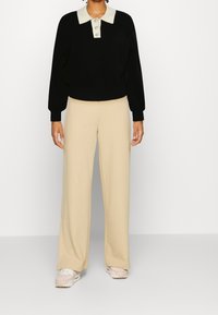 Pull noir avec col et boutons beiges, associé à un pantalon large beige et des baskets rose clair. Tissu lisse avec une coupe décontractée.