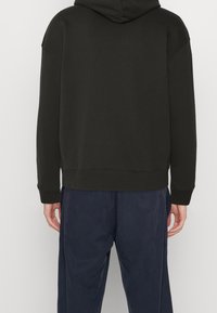 Sudadera con capucha de color verde oscuro, con un ajuste relajado, puños y dobladillo acanalados. Se lleva con pantalones azul marino, mostrando una tela lisa y texturada.