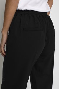 Personne portant un pantalon noir à taille élastique avec des poches arrière passepoilées et un haut blanc rentré, se tenant devant un fond uni.