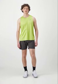 Ung mand står iført en limegrøn ærmeløs sportsbluse, sorte shorts, sorte sokker og hvide løbesko på en hvid baggrund.