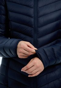 Laivasto sininen puffertakki, jossa on zigzag tikattu muotoilu, vetskari edessä ja istuvat hihansuut. Materiaali näyttää sileältä ja hieman kiiltävältä.