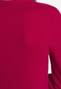 Anpassad stickad tröja i stark magenta, med en ribbad halsringning och slät yta. Bakifrån syns sömlösa axlar och rena linjer.