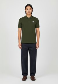 Polo shirt verde oliva de manga corta con una tapeta de tres botones, que presenta un logo blanco. Combinado con pantalones de pierna ancha en color navy y zapatos marrones.