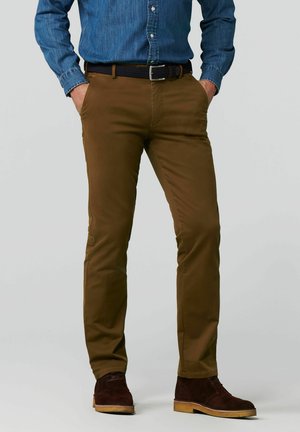 Man staat met handen in de zakken, draagt een bruine broek, een blauw denim overhemd, een donkere riem en bruine suède schoenen.