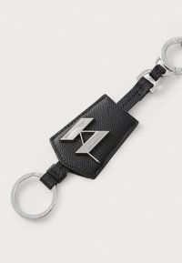 Porte-clés en cuir noir avec une finition texturée, comportant un logo en métal argenté et un anneau. Le design inclut une étiquette rectangulaire et un détail tressé.