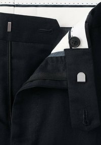 Nærbillede af linningen på sorte bukser med lynlås, knap og lukning med metalhætte samt synlig hvid og sort indvendig foring.