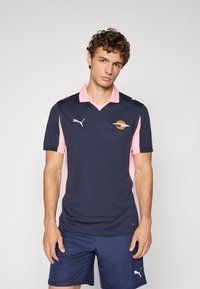 Tamsiai mėlynas ir rožinis sportinis polo marškinėlis su V formos iškirpte, trumpomis rankovėmis, Puma logotipu ir apskritu emblematu ant krūtinės. Dėvimas su tamsiai mėlynais šortais.