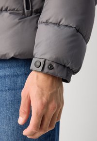 Gray puffer jacket sleeve with a smooth texture, featuring a button closure and gathered elastic cuff. Hand resting on blue denim fabric.