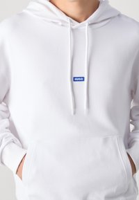 Sudadera blanca con un bolsillo frontal tipo canguro, capucha con cordón y parche azul con el logotipo "HUGO" en el pecho. Textura suave y corte regular.