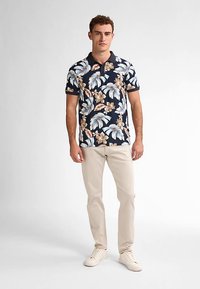 Tricou polo bleumarin cu model floral în nuanțe bej și albastru deschis, realizat dintr-un material moale, asortat cu pantaloni deschiși la culoare și adidași.