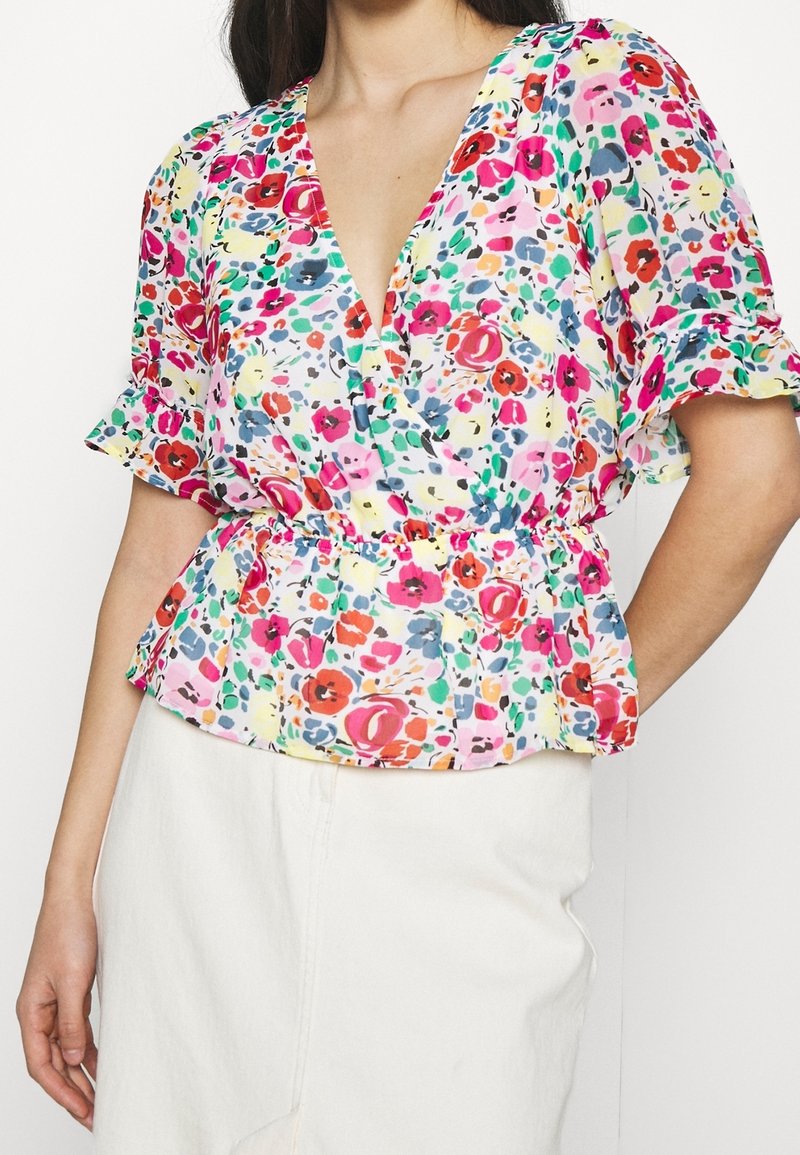Femme portant un chemisier coloré à manches courtes fleuri, avec un col en V profond et une taille élastique, associé à une jupe blanche.