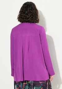 Blusa viola con una vestibilità rilassata, maniche lunghe e un dettaglio di piega sul retro, realizzata in tessuto morbido. L'orlo inferiore è leggermente curvato.