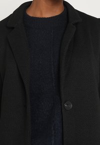 Cappotto nero con texture, colletto, chiusura a singolo bottone e maglione lavorato a maglia blu navy sottostante. Inquadratura ravvicinata che mette a fuoco i dettagli del capo.