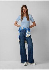 lichte blauwe top met korte mouwen, gecombineerd met hoge taille, wijduitlopende donkerdenim jeans. Gebreide blauwe en crèmekleurige trui vastgebonden in de taille. Witte sneakers.
