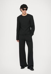 Zwarte langwerpige shirt met een ronde halslijn en een minimalistische zakdetail, gecombineerd met zwarte wijde broek en zwarte zonnebril.