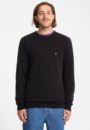 Jeune homme aux cheveux bruns mi-longs portant un pull noir à manches longues et un jean bleu, debout devant un fond blanc uni.