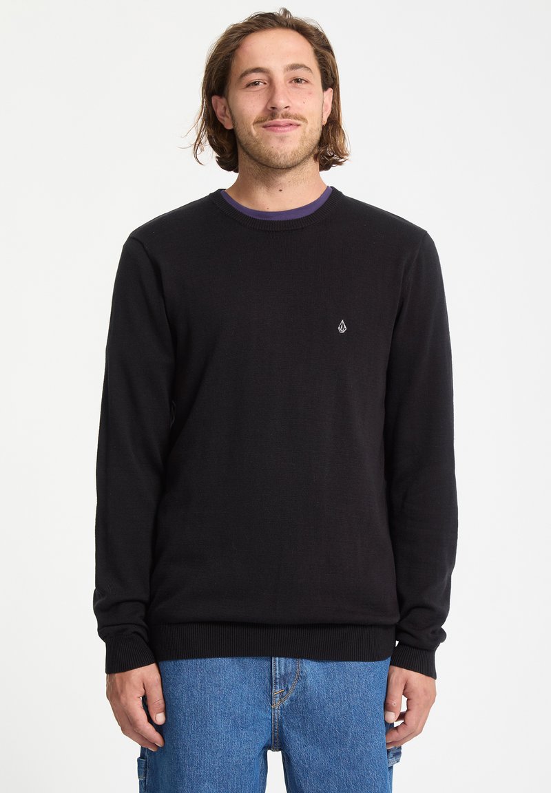 Jeune homme aux cheveux bruns mi-longs portant un pull noir à manches longues et un jean bleu, debout devant un fond blanc uni.