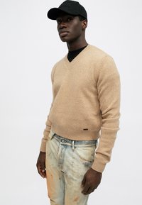 Jersey de lana beige con escote en V, con puños y dobladillo acanalados, llevado sobre una camiseta negra, combinado con jeans de mezclilla azules claros desgastados con manchas visibles.