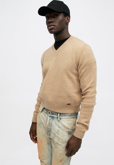 Beige Wolle-V-Ausschnitt-Pullover mit gerippten Bündchen und Saum, getragen über einem schwarzen T-Shirt, kombiniert mit verwaschenen hellblauen Jeans mit sichtbaren Flecken.