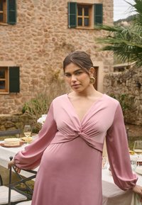 Femme en robe rose à manches longues avec un détail torsadé sur le devant, debout près d'une table extérieure dressée avec des verres à vin et un gâteau, à proximité d'un bâtiment en pierre.
