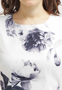 Halenka s květinovým vzorem, na bílem pozadí se nachází velké temně šedé a černé květiny. Materiál vypadá hladce a má zaoblený výstřih.