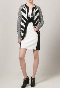 Cardigan a righe bianche e nere con apertura frontale, maniche lunghe e dettagli grigi, indossato sopra un vestito bianco aderente, abbinato a stivaletti alla caviglia.