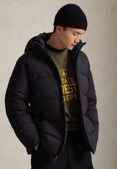 Zwarte donsjas met capuchon, met een doorgestikt ontwerp en een voorkantrits. Gedragen over een groen sweatshirt met gele tekst. Zwarte muts.