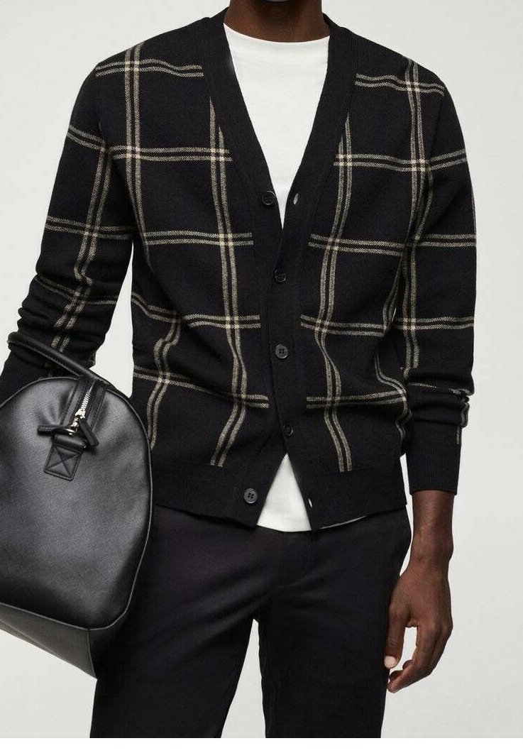 Homme portant un cardigan noir à rayures beige en plaid sur une chemise blanche, tenant un sac en cuir noir, habillé d'un pantalon noir.