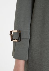 Green textured fabric with a rolled cuff secured by a square tortoiseshell and metallic clasp. Close-up of sleeve detail.