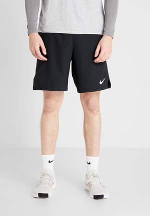 Mann trägt schwarze Nike-Shorts, weiße Nike-Socken und hellgraue Sneaker, steht vor einem einfarbigen hellen Hintergrund.